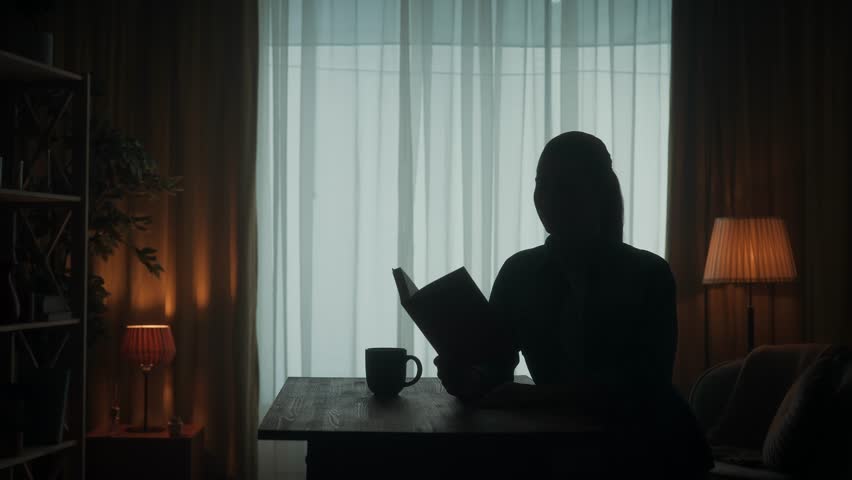 A woman reads a book sitting at a table with a cup of coffee, tea in the room. Dark silhouette of a woman with an open book in her hand, reading, turning pages, drinking coffee. Slow motion.