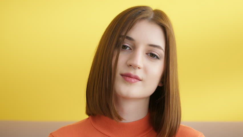 Portrait of positive smiling young woman with square haircut on yellow background. Attractive brunette looks at camera, angling her head left and right. High quality 4k footage Royalty-Free Stock Footage #1105918247