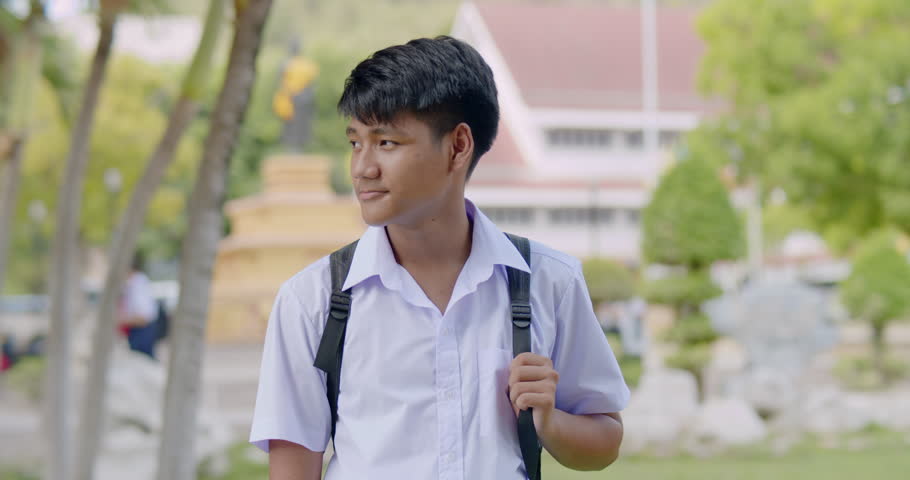 Slow motion of A happy smiling Asian male high school student in white uniform with backpack waiting for car to go back home after school in the evening.