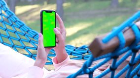 A woman is holding a phone with a chromakey screen and swinging in a hammock in a green garden.