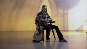 Young musician with long hair is sitting in an armchair with a guitar standing next to him.