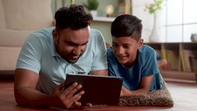 joyful indian father with son using digital tablet while lying on floor at home - concept of weekend holiday, Modern parenting and Digital entertainment