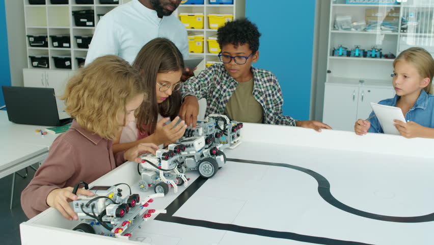 Medium shot of excited nine-year-old diverse children testing their electric robot models at kids hobby club and pressing buttons on control panels, and black male teacher watching and guiding Royalty-Free Stock Footage #1105936159