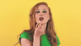 Pleasant red-haired woman puts on makeup in front of the camera. A young woman stands on an orange background and paints her lips.