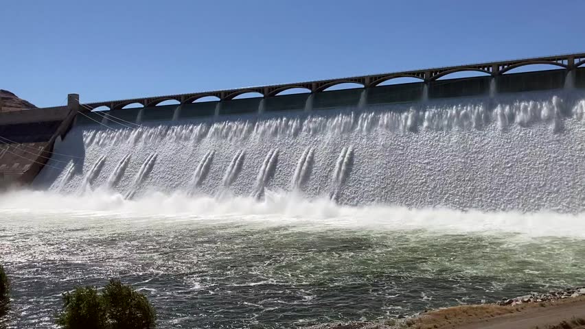 Tracking shot of the full length of Grand Coulee Dam with water flowing over the spillway at peak Spring flow. It's unusual to see this amount of water. Taken on a sunny day - Eastern Washington, USA Royalty-Free Stock Footage #1105943497