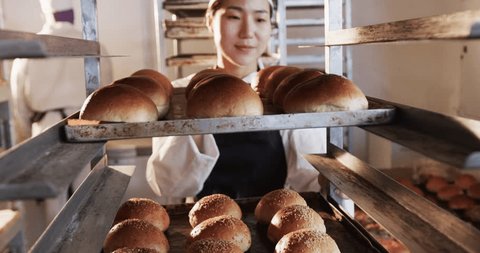 Happy asian female baker working in bakery kitchen, holding baking sheet with rolls in slow motion. Food, small business, baking, bakery and work, unaltered. 스톡 비디오