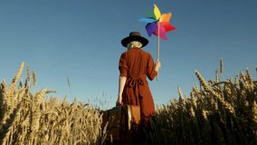 Stylish woman in brown vintage dress and hat with retro suitcase and pinwheel in golden wheat field