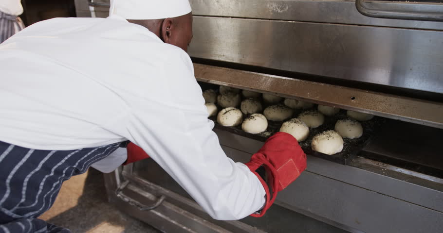 African american male baker working in bakery kitchen, putting rolls into oven in slow motion. Food, small business, baking, bakery and work, unaltered. Royalty-Free Stock Footage #1105947907