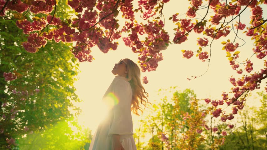 Happy young woman spinning dancing enjoy spring nature green leaves trees pink sakura flowers sun light day park. Beauty cheerful girl smiling face, long blond hair flying in wind motion. White dress