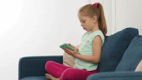 Happy little girl with a tablet in her hands sits on the sofa at home. Cute teenage child watching cartoon or playing game on digital tablet at home. Children's leisure of the modern child. 
