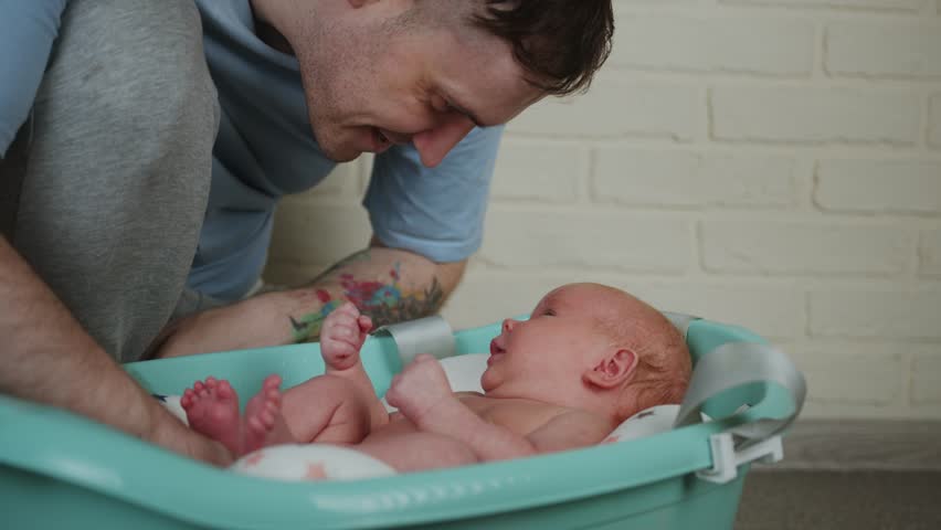 Dad with tattoos bathes his newborn. Bathing a newborn baby in a bathtub. A lovely child is undergoing military training for the first time.