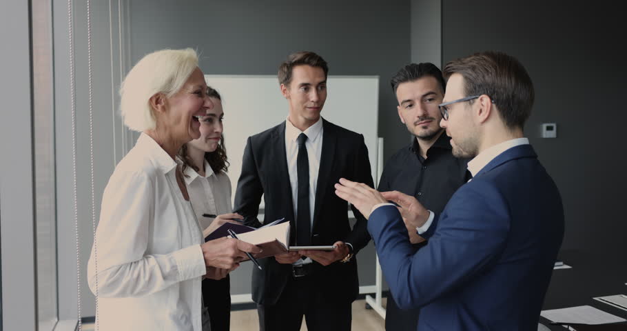 Positive businessman in formal clothes telling funny story to colleagues on work break. Group of coworkers standing together in workspace hall, talking, laughing, having fun, enjoying leisure Royalty-Free Stock Footage #1105962957
