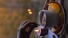 Woman hand mixing tea with citrus fruits with spoon, fruits whirling in the cup, candle goes out. Cozy home autumn background. Having cup of tea near the window. Vertical video 