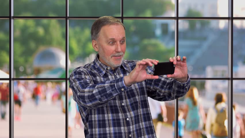 Happy smiling cheerful senior man takes a photo on his smartphone camera. Checkered indoor window with city street view.