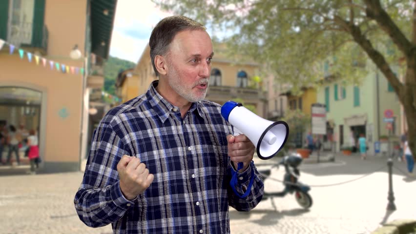 Mature man standing outdoors giving a speech in megaphone and gesturing. Blurred european town life in the background.
