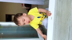Lovely little boy sitting at the door to a balcony. Cute child wearing yellow t-shirt at home. Vertical video.