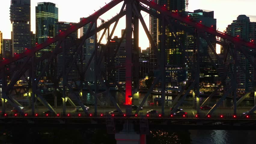 Story bridge adventure climb, tiny people climbing on iconic cantilever bridge with busy traffic crossing the river at dusk and illuminated downtown cityscape of Brisbane city, cinematic aerial shot.