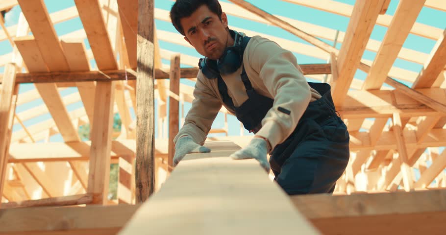 Builder man holding wooden plank, pulling it, strong man carrying board. Attractive guy in uniform builds house out of wood. Wooden house frame. Construction loader worker. Royalty-Free Stock Footage #1105983913