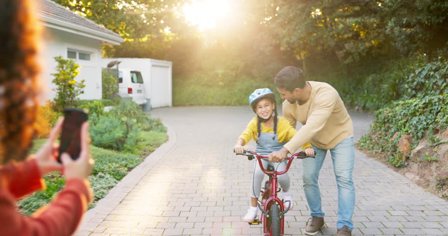 Happy family, parents and teaching kid on bicycle with phone to record video, photograph and childhood growth. Mom, dad and memory of girl learning to ride bike with support, play and development