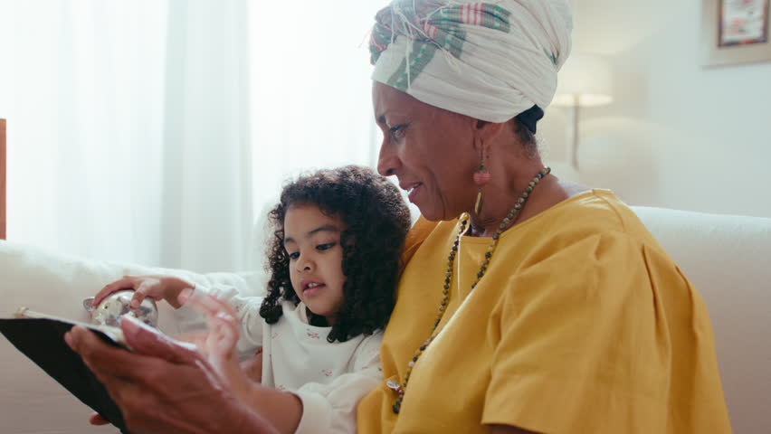 Senior black woman sitting on sofa, embracing little granddaughter, using digital tablet and talking to child during the day at home - Powered by Shutterstock - Get 15% off with code: PIKWIZARD15