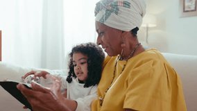 Senior black woman sitting on sofa, embracing little granddaughter, using digital tablet and talking to child during the day at home - Powered by Shutterstock - Get 15% off with code: PIKWIZARD15