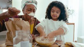 Happy senior African-American grandmother teaching little girl to bake, putting flour into bowl and talking, kid mixing ingredients for dough at kitchen table at home - Powered by Shutterstock - Get 15% off with code: PIKWIZARD15