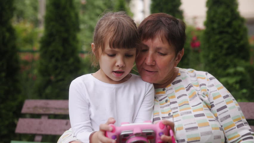 Cute girl granddaughter shows her grandmother photos on her toy camera. The family is sitting on a bench viewing photos on a camera. Different generations are spending time together. Royalty-Free Stock Footage #1105998137