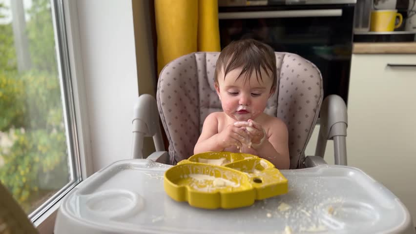 baby girl eats dirty. happy family child dream concept. baby funny got dirty in yogurt lifestyle dairy product all over his face in drops learns to eat. baby eats dirty at home at feeding table