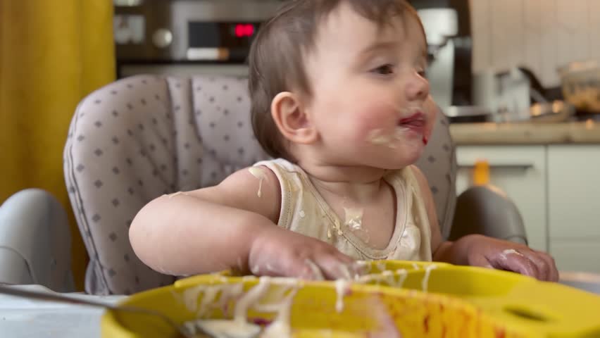 baby girl eats dirty. happy family kid dream concept. baby funny got dirty in yogurt dairy product all over his face in drops learns to eat. baby eats dirty at home lifestyle at the feeding table Royalty-Free Stock Footage #1106006075