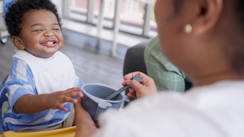 Asian mother feeding her 9 months old her cute little baby and African American helping for holding food plate At Home. Photo series of family, kids and happy people concept. Parents feed kids. Royalty-Free Stock Footage #1106006385