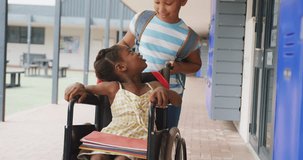 Video of two happy diverse schoolgirls, one pushing other in wheelchair in corridor, with copy space. Education, childhood, inclusivity, elementary school and learning concept.