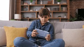 Teens and gaming addiction. Young boy playing video game on smartphone sitting on sofa at home. Cute teenager losing in car racing game on mobile phone. Spending time at home, leisure, relax.