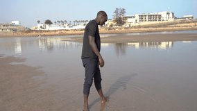 Video portrait of a male person of color in his thirties as playing with the water as he gazes upon and enjoys the beauty of the beach on a sunny day in Morocco, North Africa.