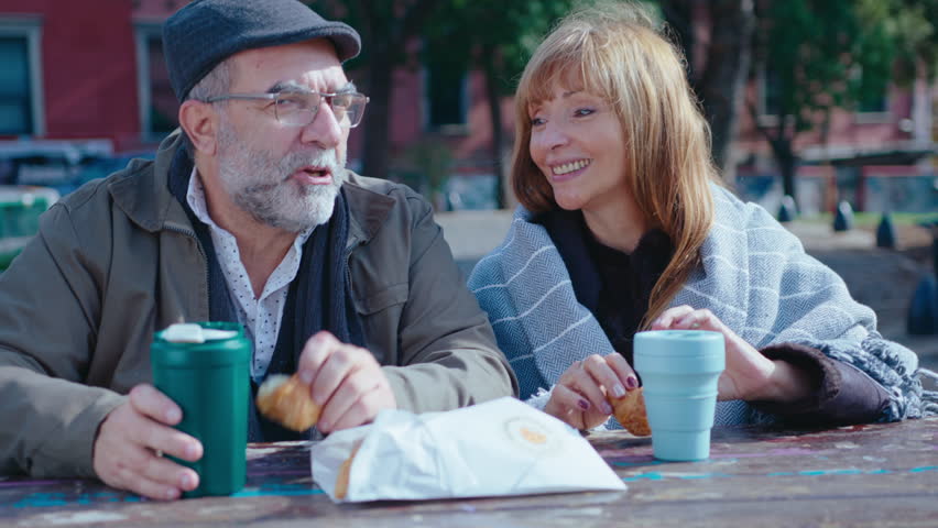 Beautiful senior woman smiling and talking with husband, sitting outdoors at table in the park, drinking coffee from travel mugs and eating pastry