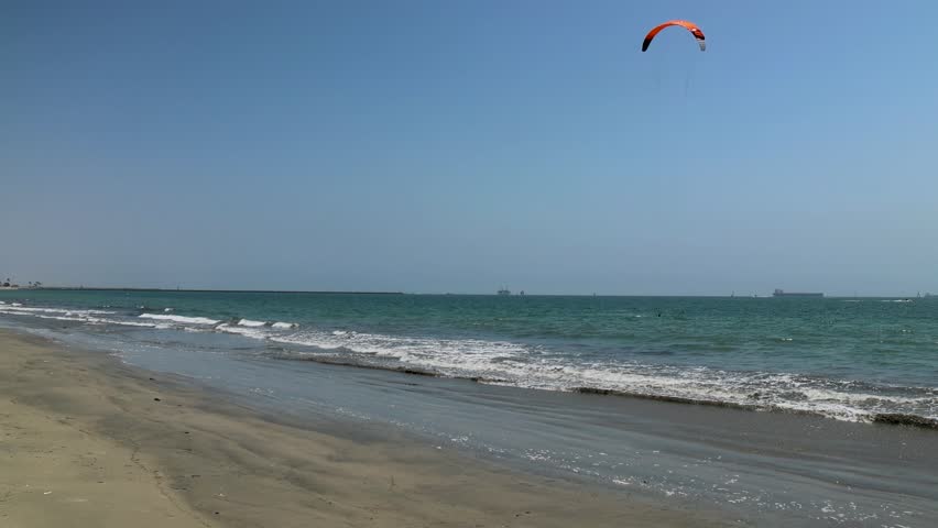 footage of a man parasailing with an orange and black parasail with motor boats and sailboats sailing across the blue ocean water Rosie’s dog beach in Long Beach California USA	