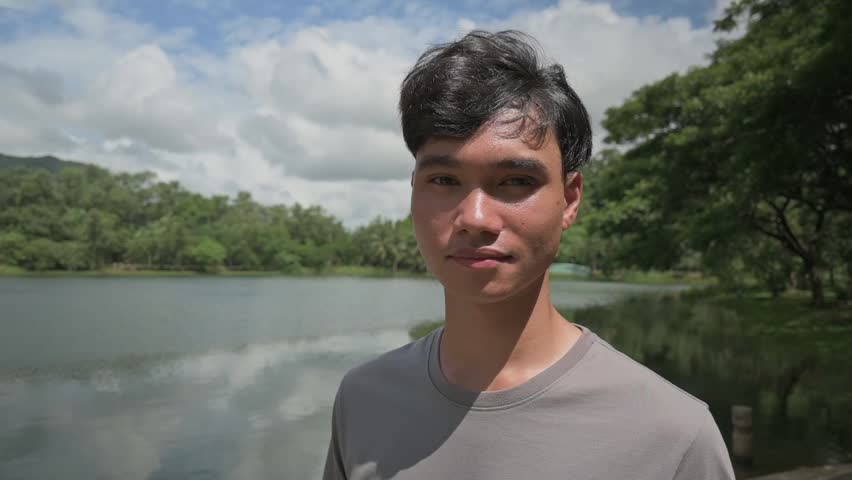 Portrait asian teen handsome boy smiling and looking at camera while standing at natural lake in public park. Young teenager lifestyle. Outdoor pursuit.