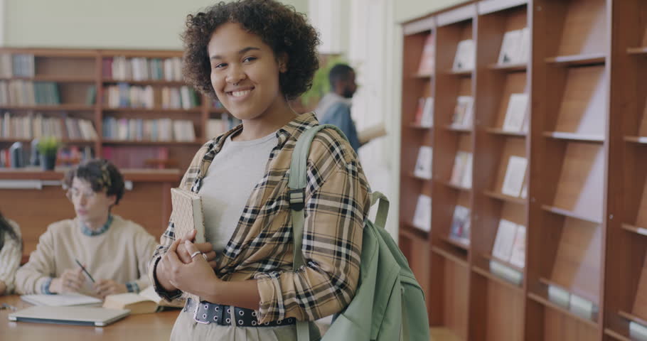 Slow motion portrait of cheerful African American woman student standing in library with books and backpack smiling looking at camera