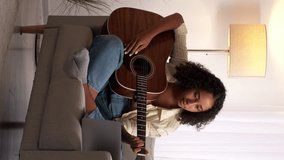 Vertical video. Music instrument practice. Guitar course. Cheerful woman guitarist studying online internet lesson on laptop sitting couch home room.