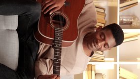 Vertical video. Guitar practice. Musician leisure. Creative hobby. Talented cheerful man playing song chords on string instrument on couch in living room at home.