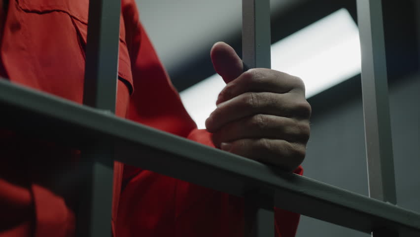 Close up of hands of prisoner in orange uniform holding metal bars, standing in prison cell. Guilty criminal serves imprisonment term for crime. Inmate in jail, correctional facility. Justice system. Royalty-Free Stock Footage #1106034963