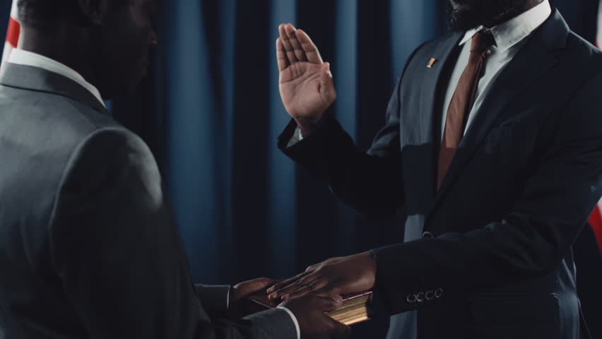 Medium tilting shot of bearded middle-aged bearded black male politician in suit, tie and glasses giving oath on holy Bible or Quran, with raised hand, American flag and dark blue drapes in background Royalty-Free Stock Footage #1106039341