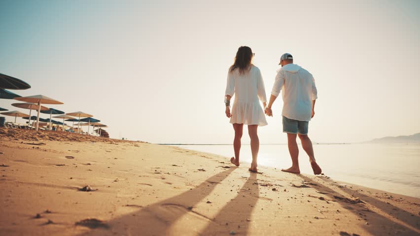 Loving couple walking along sandy ocean beach at sunny day talking enjoying time together. Man woman having romantic date, honeymoon near sea, back view. Tourism, travel family holidays concept. Royalty-Free Stock Footage #1106040389