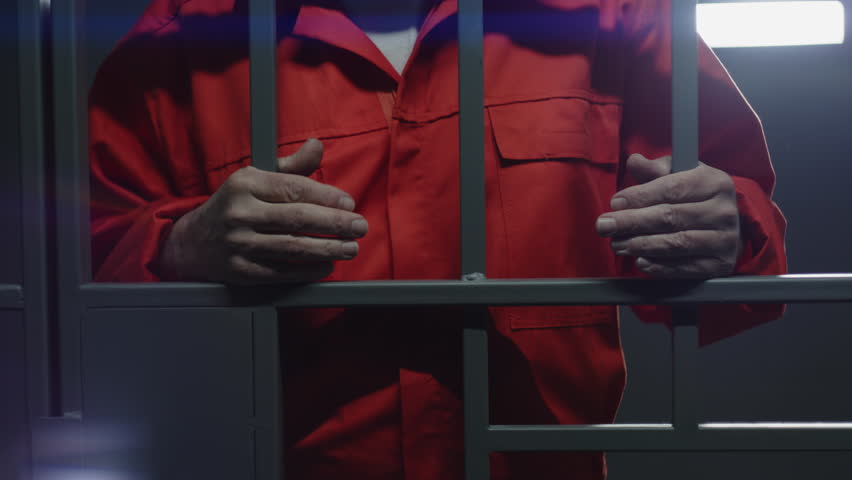 Close up of prisoner in orange uniform holding metal bars, standing in prison cell. Criminal or murderer serves imprisonment term for crime. Inmate in jail, detention center or correctional facility. Royalty-Free Stock Footage #1106040903