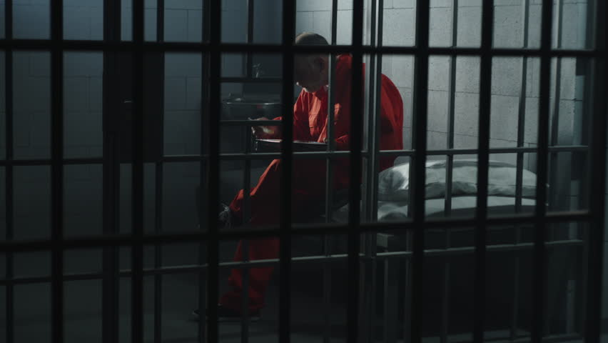 Elderly prisoner in orange uniform sits in prison cell, eats food. Criminal serves imprisonment term for crime. Conditions in jail or correctional facility. Shooting from jail cell through metal bars. Royalty-Free Stock Footage #1106041001