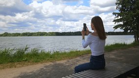 Woman processes video on smartphone to send it to messenger while sitting on bench on river bank
