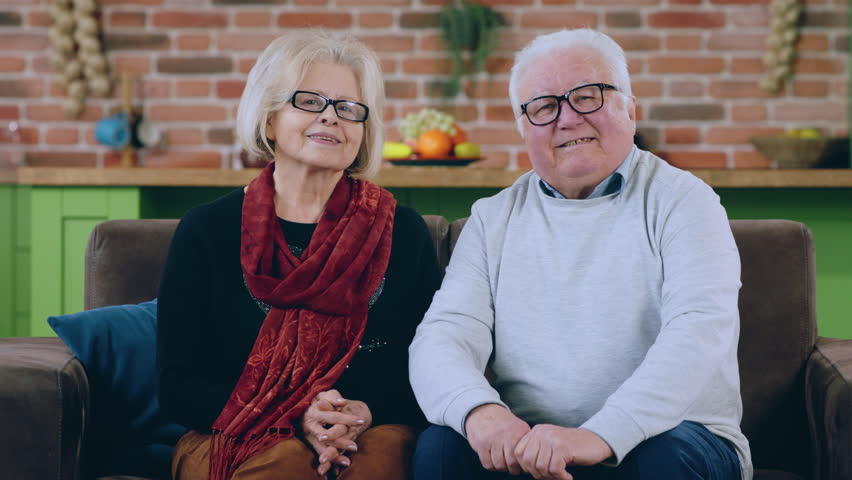 Capturing video in front of the camera looking straight and posing old couple while sitting on the sofa in living room