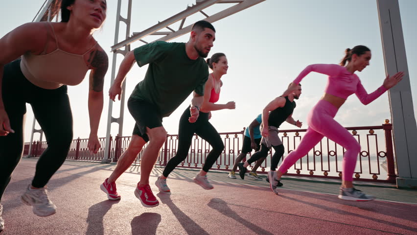 Male and female runners stand in row preparing to run. Young athletes smile talking cheerfully and run together on bridge under blue sky low angle shot
