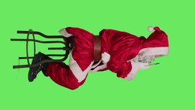 Vertical video Profile of character in red costume sit on chair, thinking about new ideas over full body greenscreen background. Young pensive man acting like santa in suit and white beard