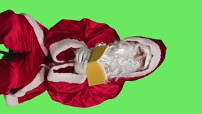 Vertical video Front view of santa reads literature sitting on chair, man wearing festive red costume while he is reading poetry book over greenscreen backdrop. Father christmas enjoying fairytale