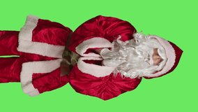 Vertical video Front view of faithful santa claus praying in studio, holding hands in a prayer and standing against greenscreen backdrop. Model portraying father christmas with festive red costume
